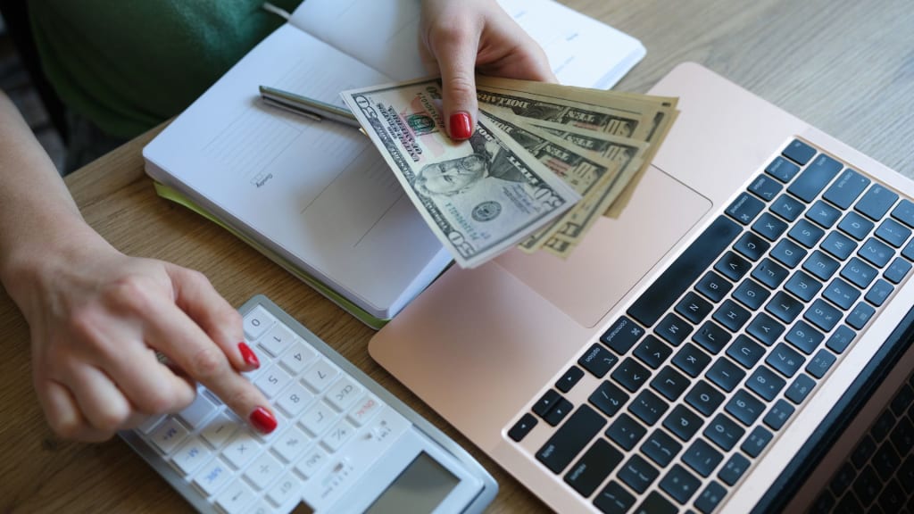 Person holding dollar bills, using a calculator, and working on a laptop at a desk.